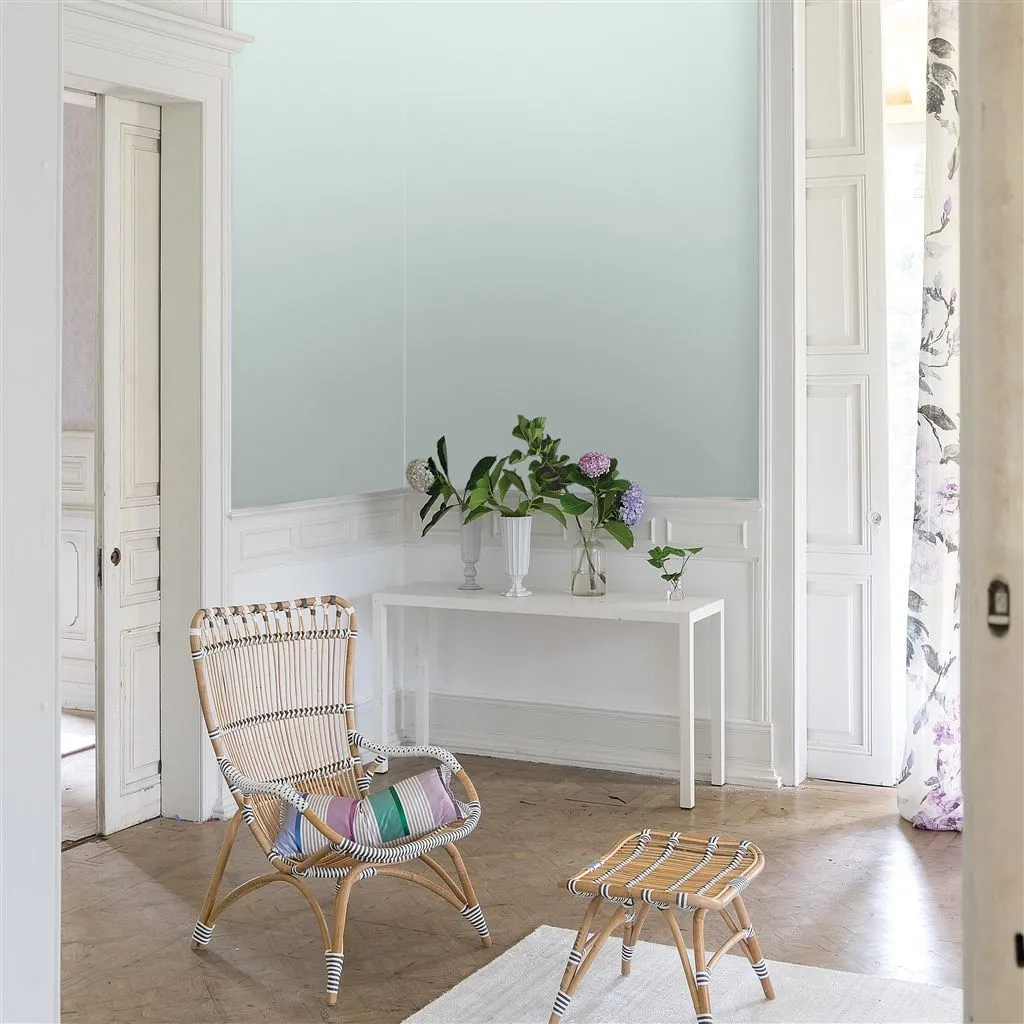 Corner of a living room painted in eau de nil by Designers Guild. Wood work is painted in white. A small sideboard with a plant stand against the wall and a rattan chair with matching footstool