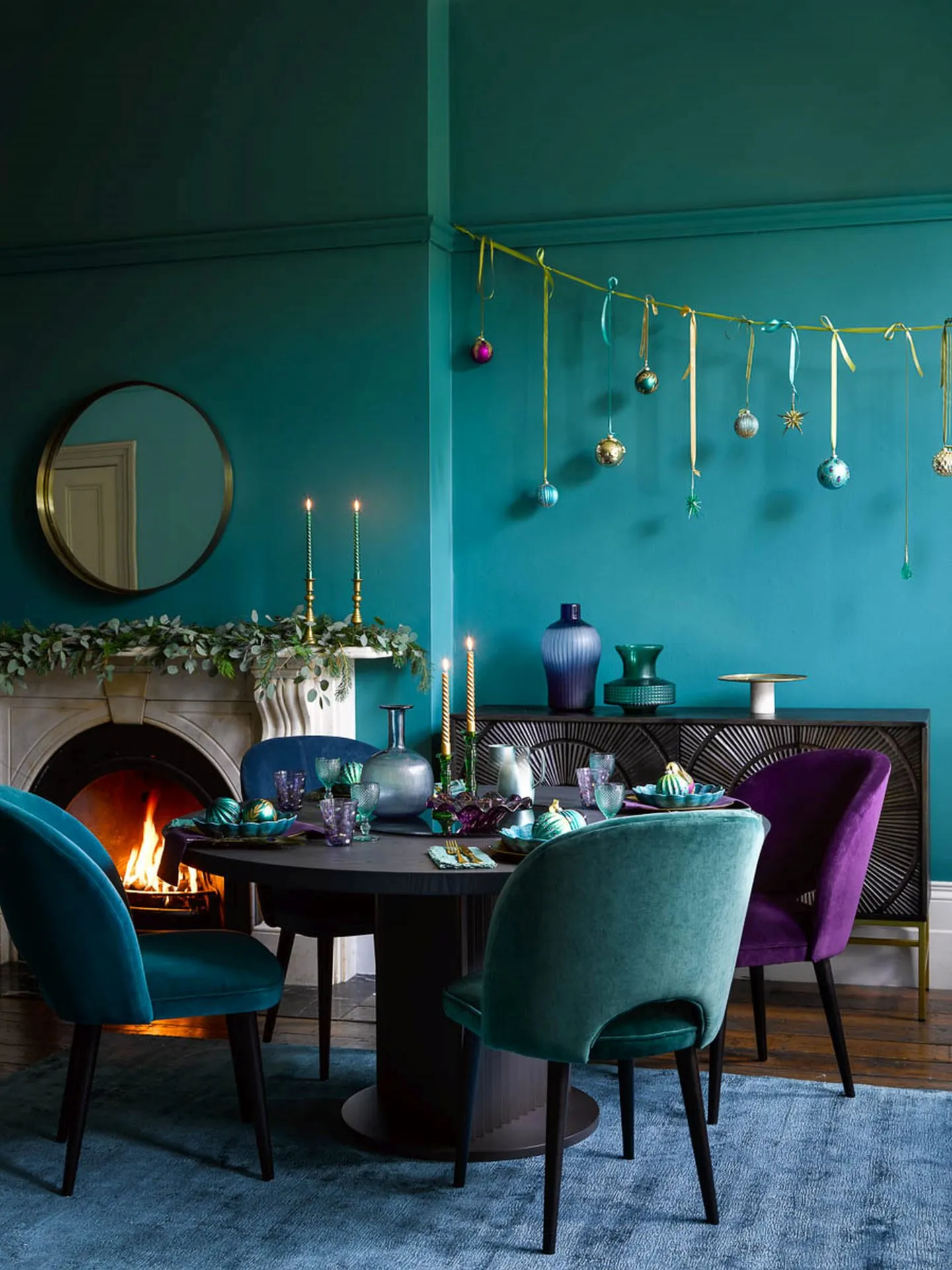 Opulent christmas tablescape in blue, green, purple, and teal with gold accents and velvet dining chairs.