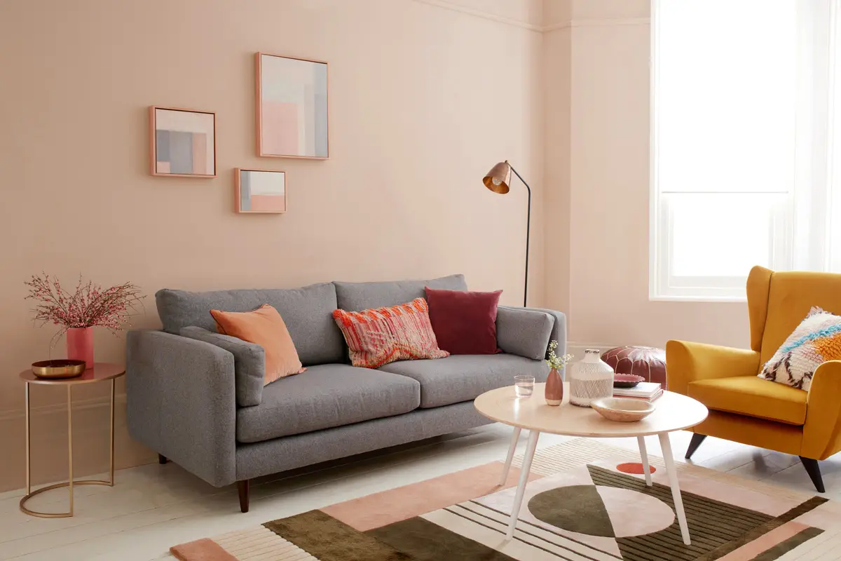 Living room with light pink wall colour, white painted wooden floors, and a grey sofa. Colourful accessories and a yellow armchair.